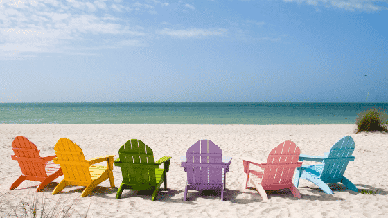 6 Adirondack chairs in a semicircle on the beach, facing the ocean and blue sky. From left to right, orange, yellow, green, lavender, pink, light blue chairs.