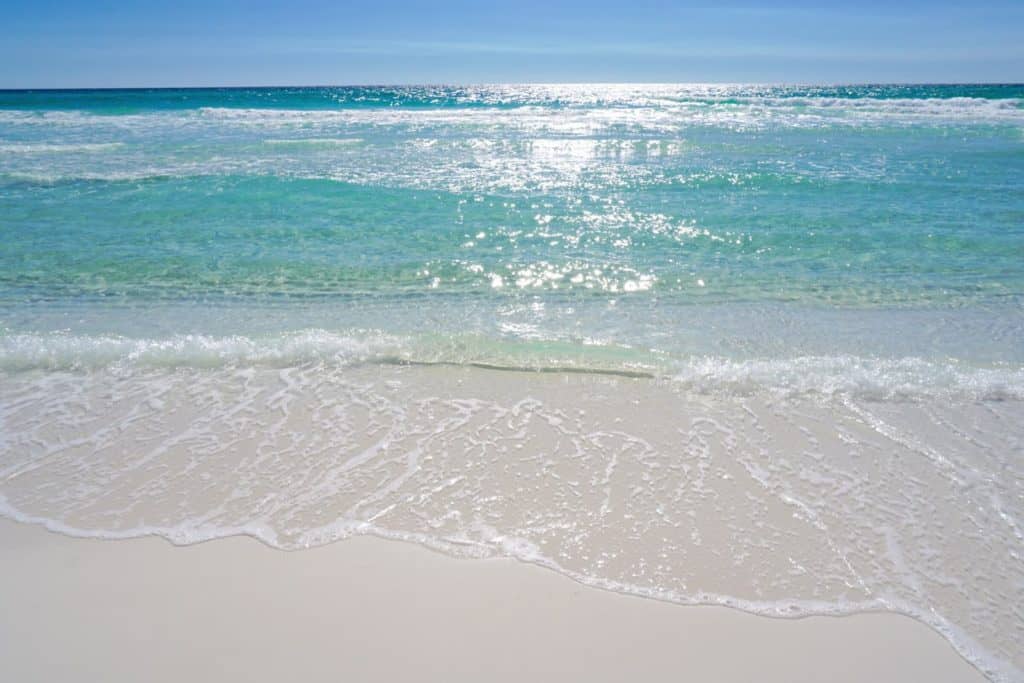 The sun is reflecting off of the clear blue and green water on Destin Beach. The ocean blends into a blue sky and water covers the white sand