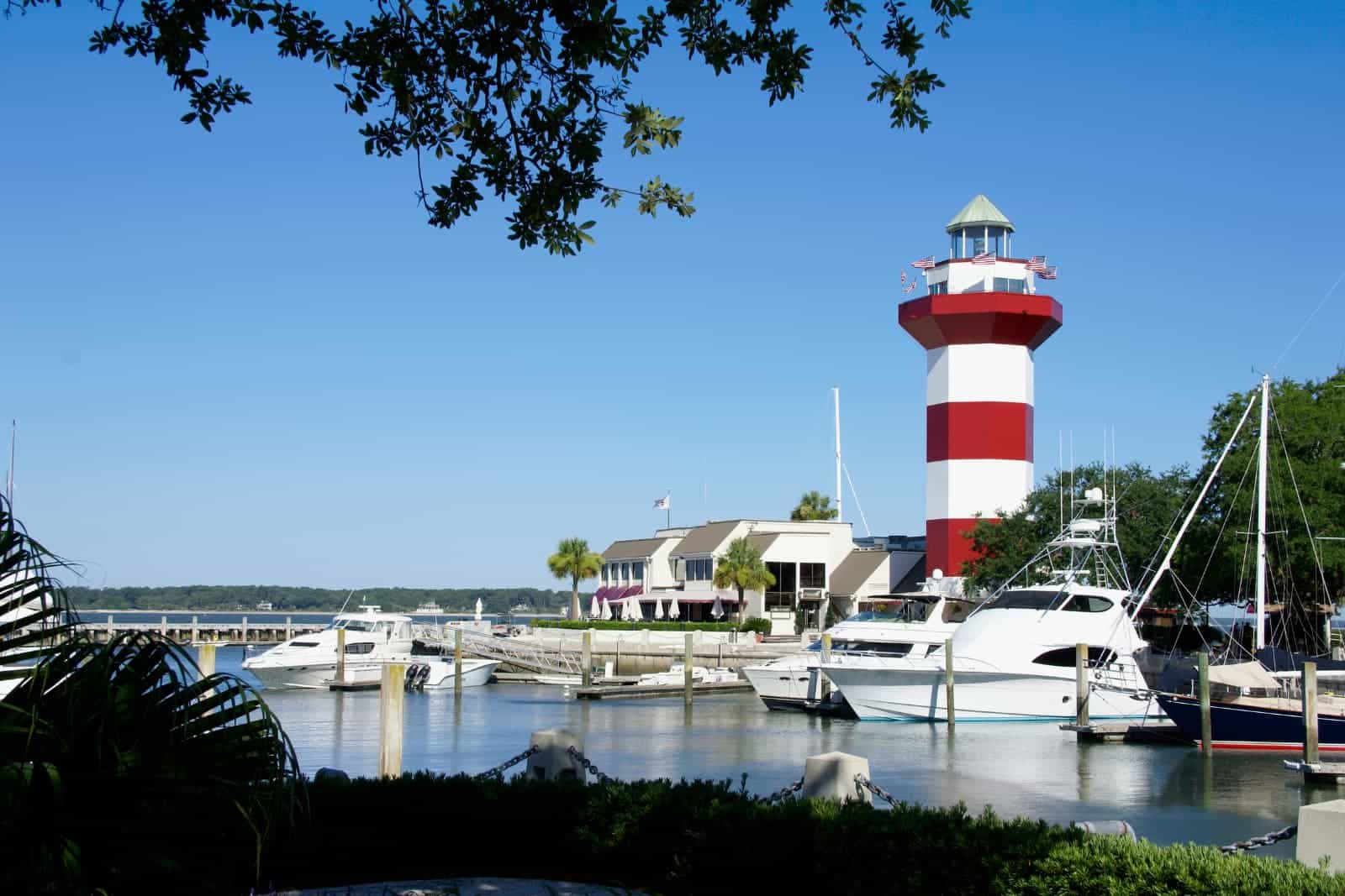 Harbour Town Lighthouse on Hilton Head Island, South Carolina. The red and white striped lighthouse is against a blue, cloudless sky and it towers above sailboats and yachts in clear blue water..