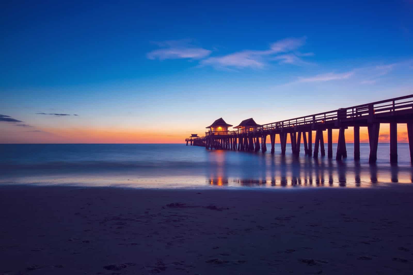 Naples Pier at sunset. A brilliant blue sky fades into orange and pink where it meets the dark blue water of the ocean. The beach is in the foreground and the pier stretches out into the ocean on the right side of the image.