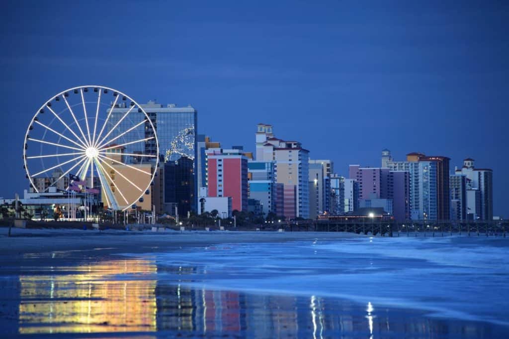 Nighttime shot of the Skywheel, a huge observation wheel, lit up with white lights. Hotels and buildings of various sizes line the beach behind the Skywheel.