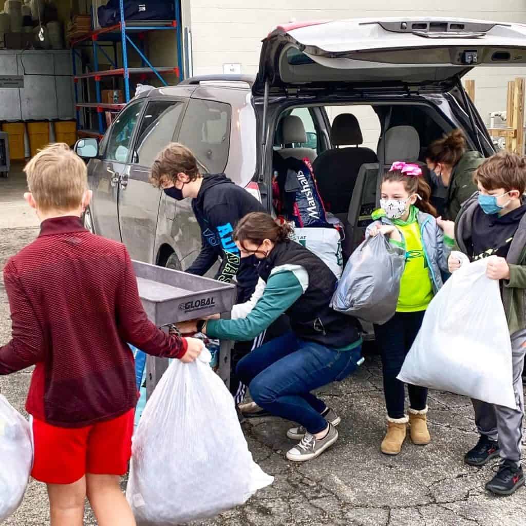 A woman and four tweens and teens unloading a van, carrying white garbage bags full of hoodies.