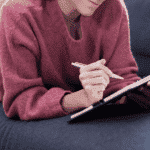 Woman in burgundy sweater on her stomach on a gray couch writing in a gratitude journal