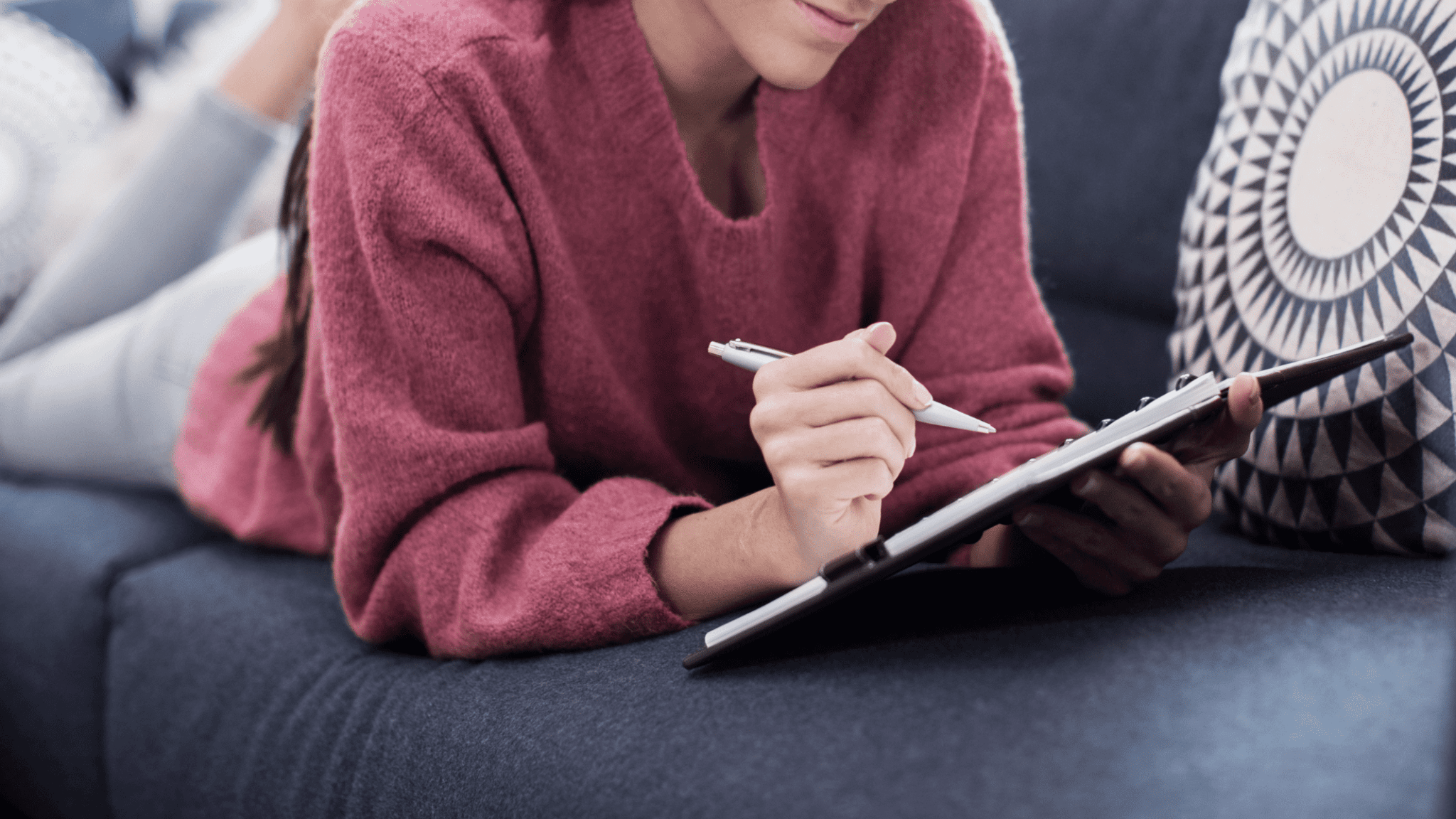 Woman in burgundy sweater on her stomach on a gray couch writing in a gratitude journal
