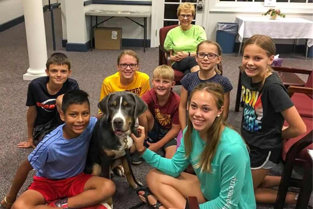 Seven tweens and teens plus one senior woman sitting on the floor of a church with a Bernese mountain dog therapy dog.