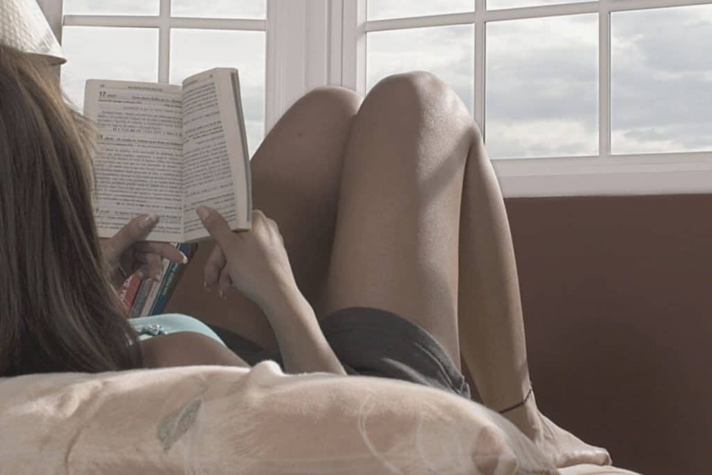 a teen girl sitting in a bed with her knees up reading a book with windows in front of her