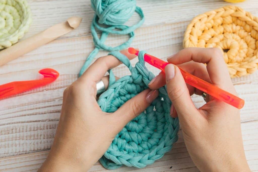 hands crocheting a blue craft with yellow and green projects and extra crochet hooks in the background