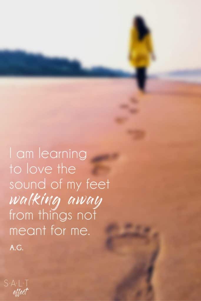 "I am learning to love the sound of my feet walking away from things not meant for me." A fresh start quote by A.G. in white text. Background is a blurred image of a woman walking on the beach in a yellow jacket. Her footprints are visible in the sand.