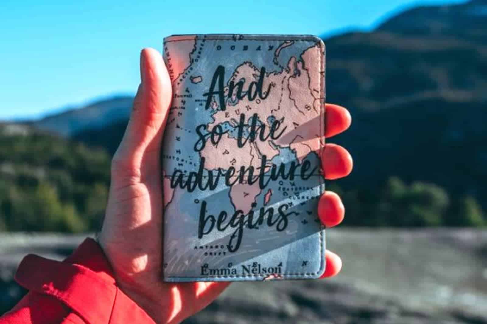Woman's hand holding a passport cover with a world map on it and the words "And so the adventure begins." A mountain range is blurred in the background
