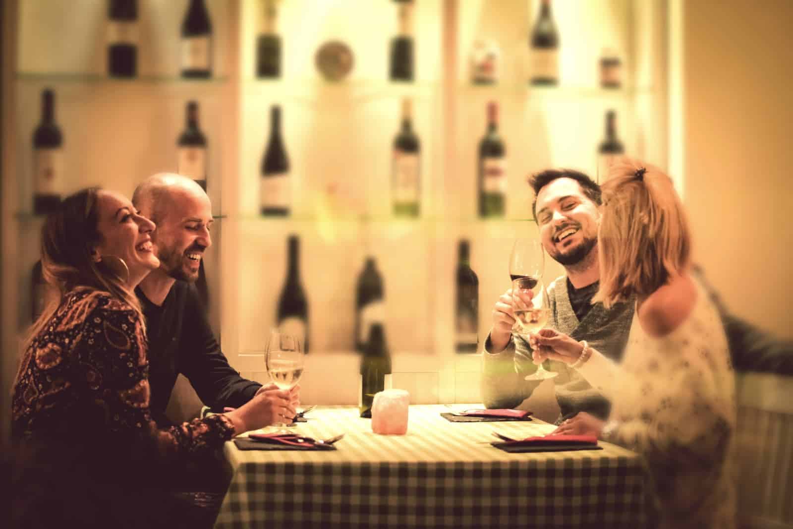 Two couples laughing at a table in a restaurant. All are holding wine glasses and the wall in the background is full of shelves of wine.