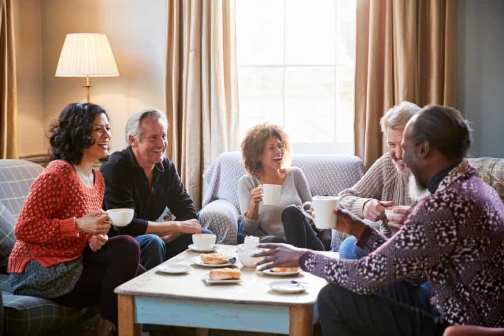 Five middle age adults sitting around a table on couches in a living room. They are laughing together as if they've been couple friends for a long time.