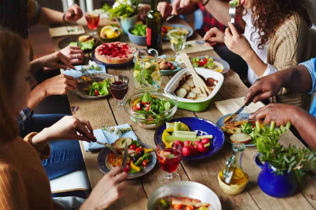 Colorful platters of food on a long wooden table. Couple friends are sitting around the table eating salads, vegetables, bread and potatoes.