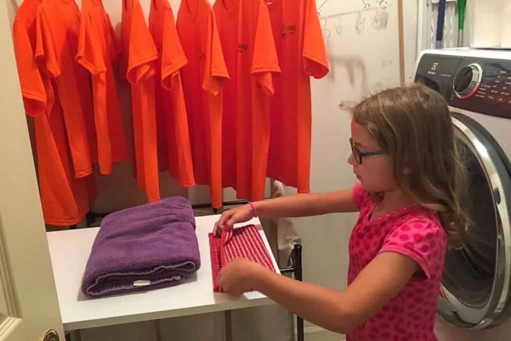 A girl in a pink t-shirt and glasses folds laundry next to a dryer and a rack of hanging orange tshirts.