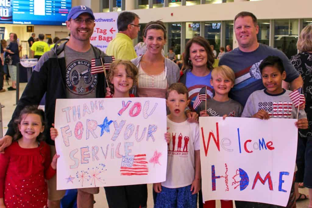 Two families (four adults and five tweens) are dressed in red, white and blue. They hold patriotic signs and flags in an airport.