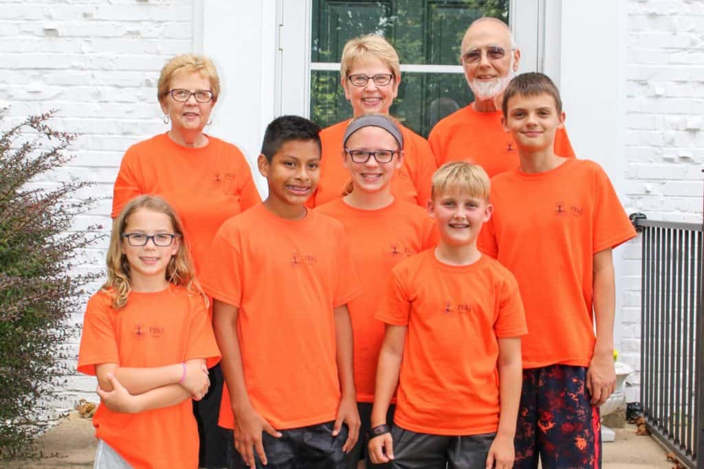 Five tween and teen kids standing on steps with three senior adults. All are wearing matching orange tshirts with the PB&J Camp logo.