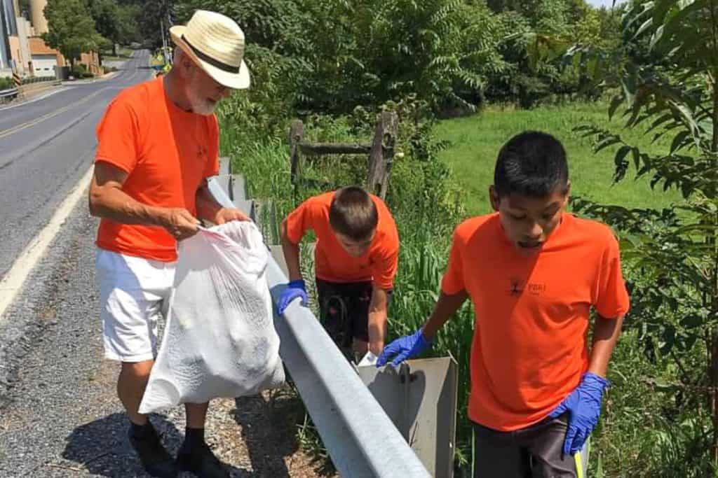 Two young teenage boys and a senior man in matching orange tshirts are picking up litter in a grassy area behind a guard railing.