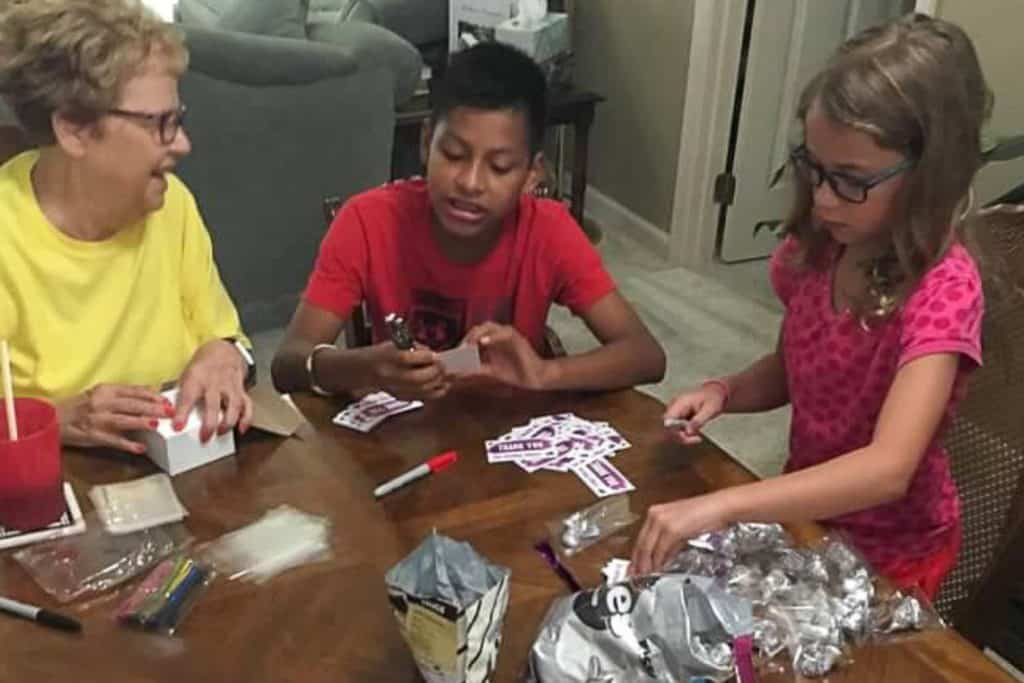 A boy, girl, and grandma sitting at a table. They are putting Hershey kisses in baggies and adding tags that say "thank you for serving others."