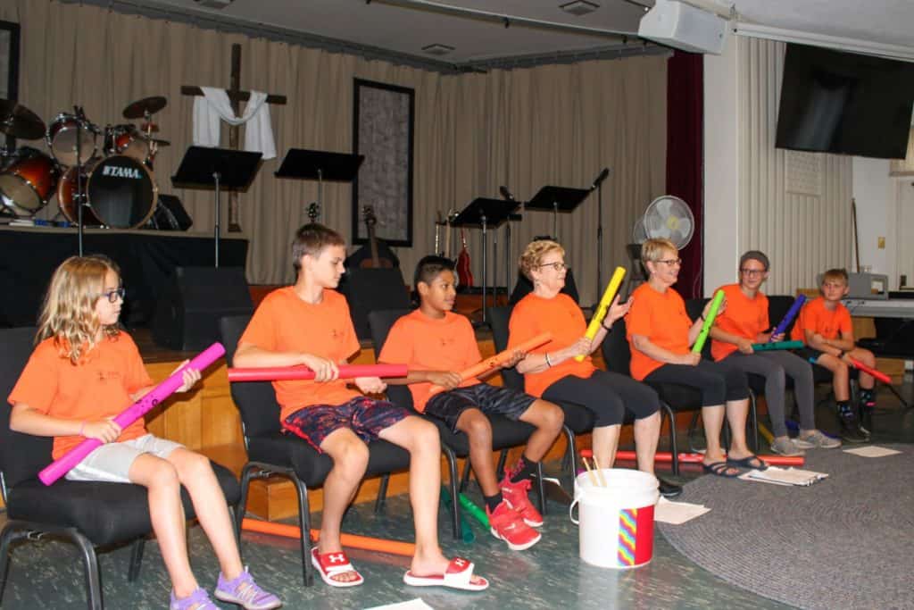 Five tweens and teens plus two senior women are dressed in matching orange tshirts. They are using instruments as part of their volunteer talent show at a nursing home.