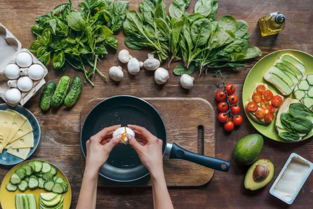 a table covered in fresh produce and hands breaking an egg over a frying pan