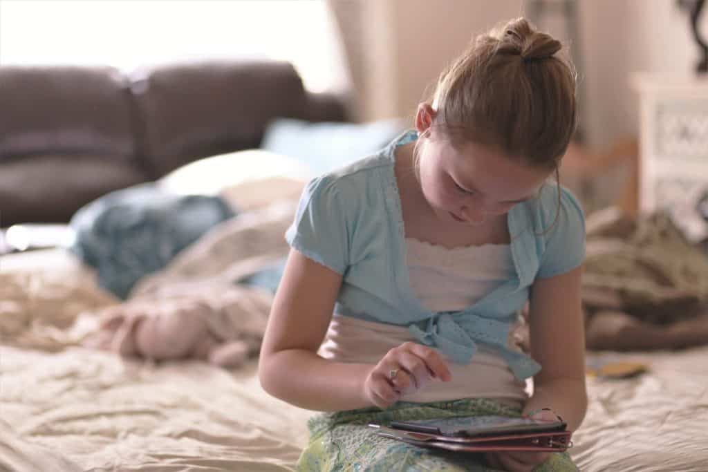 Tween girl sitting on the edge of an unmade bed using her tablet in her lap.