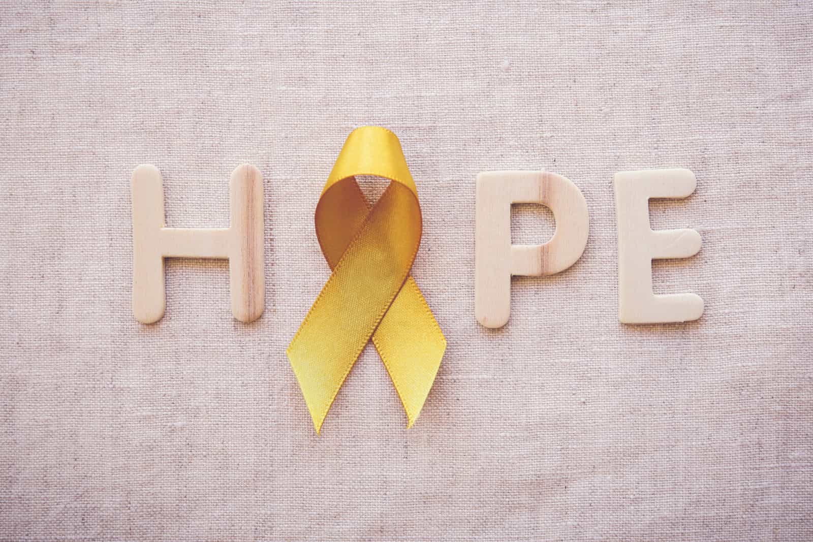 HOPE in light wooden letters on a cream linen background. The "O" is a gold ribbon for Childhood Cancer Awareness Month