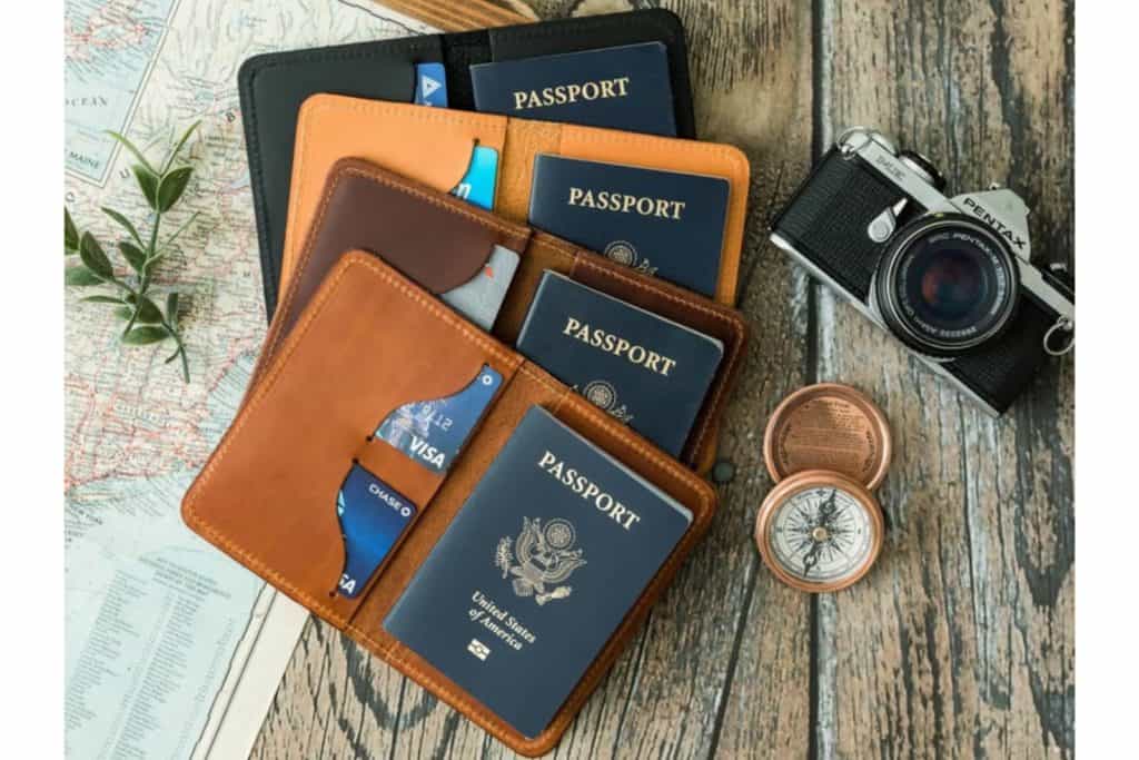 Stack of 4 leather passport holders on top of a map and next to a compass and camera. Passports are included and the covers are in 3 shades of brown and black.