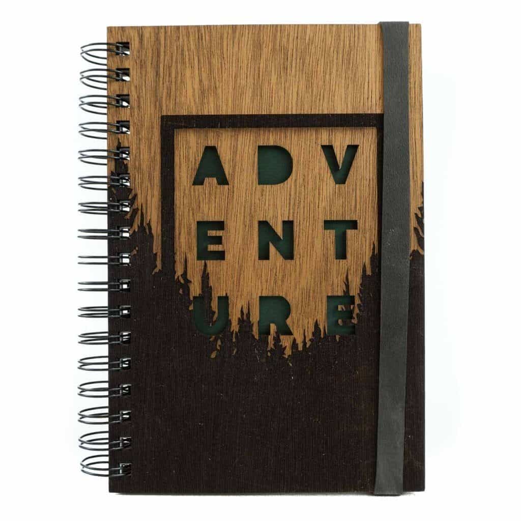 Spiral bound wooden journal with a burnout image of pine trees and the word "Adventure" in all capital letters