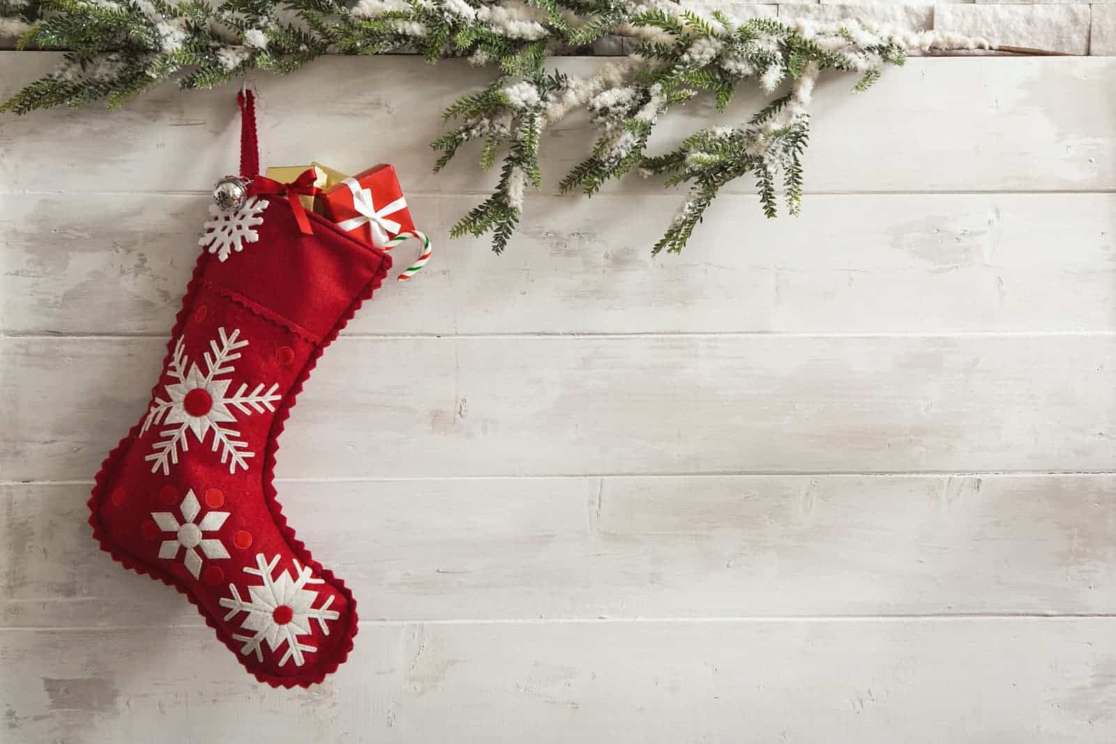 Red stocking with white snowflakes hanging against a white wood wall with pine branches covered in snow.