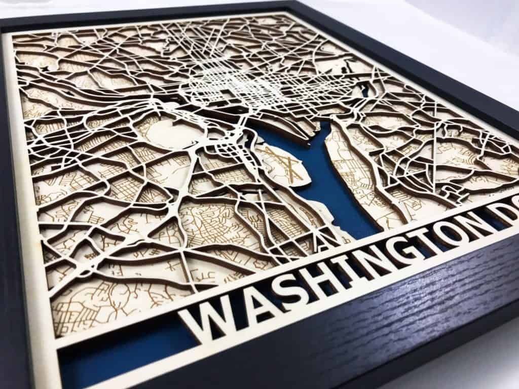 3D laser cut wood map of Washington DC or college town. Black frame, carved light wood and blue painted area for water.