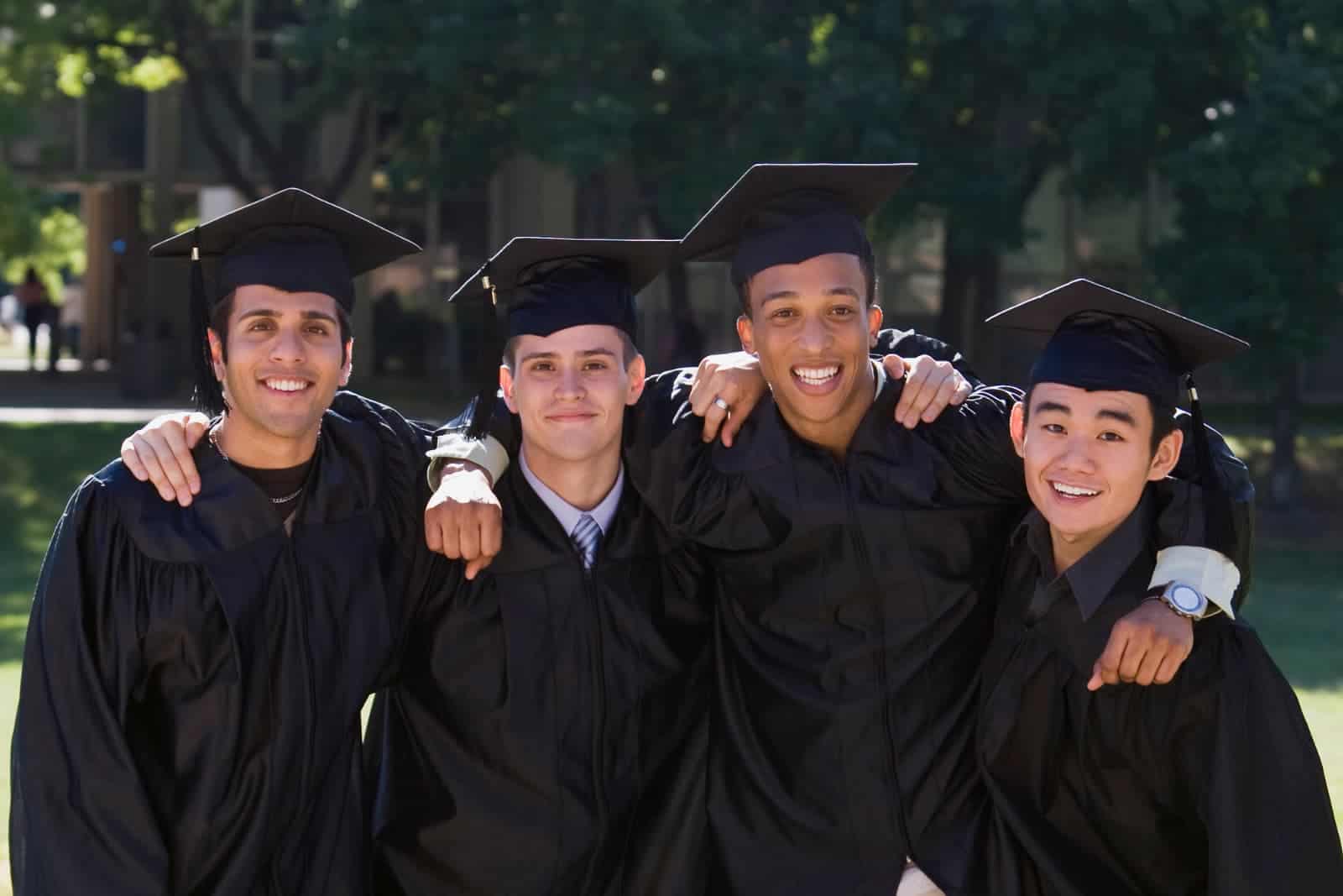 Four college guys wearing caps and gowns at their college graduation. Smiling photo with arms around each other.