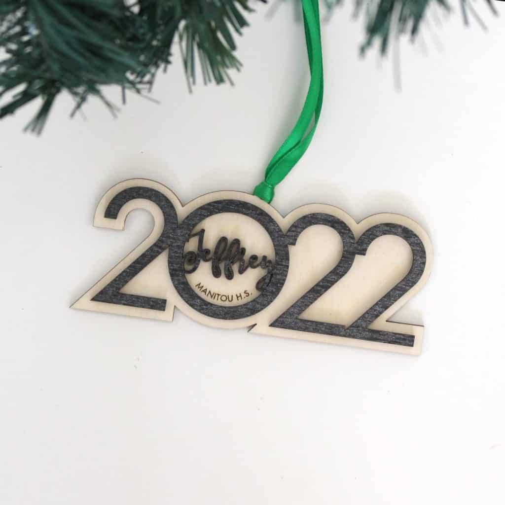 Class of 2022 ornament best college graduation gifts. Light wood background and gray wood carved 2022 with Jeffrey and Manitou H.S. inside the 0. Green ribbon hanging on tree.