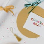 Flay lay on a white table with colorful metallic glitter circles. Diploma tied with gold ribbon, gold tassel, gold circle behind a card that says "congrats grad" in colorful letters.