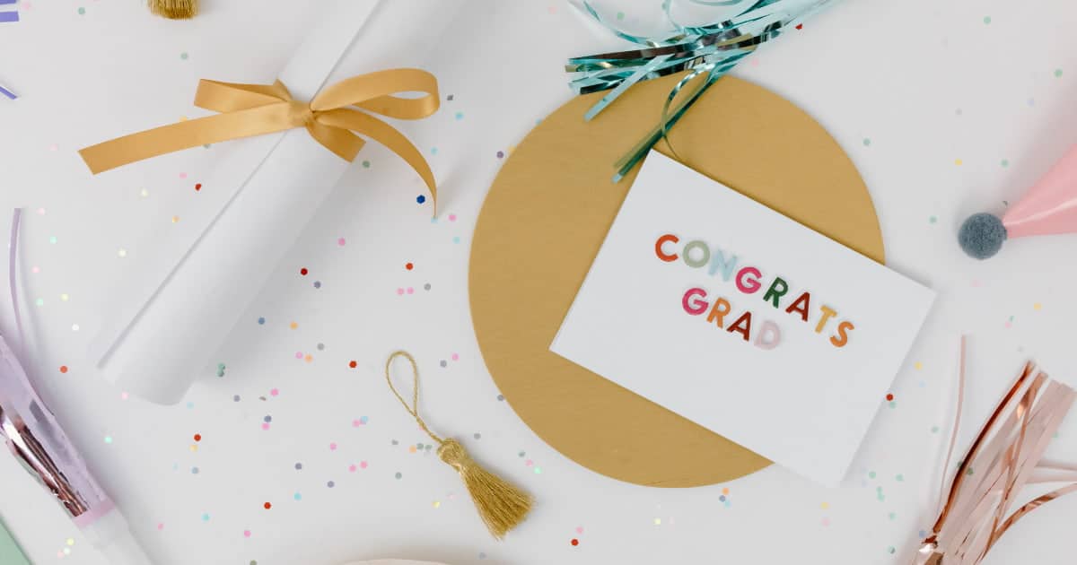 Flay lay on a white table with colorful metallic glitter circles. Diploma tied with gold ribbon, gold tassel, gold circle behind a card that says "congrats grad" in colorful letters.
