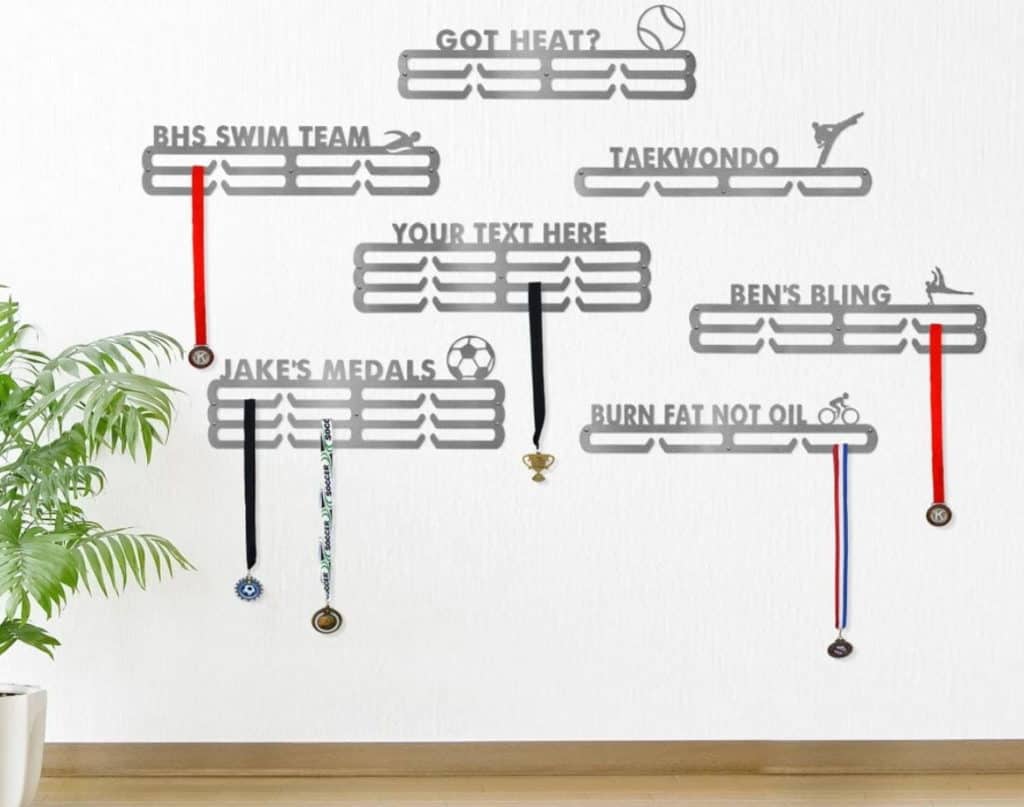 7 medal racks hanging on a white wall. Racks are silver and have 1, 2, or 3 rows for hanging medals and custom text and icons on top: "Got heat?" with a baseball; "BHS swim team" with a swimmer; "Taekwondo and a martial arts competitor