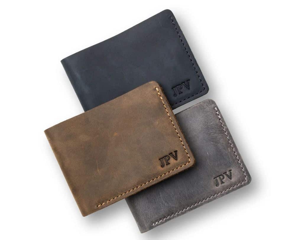 3 stacked distressed leather wallets with initials stamped on bottom right corner. Navy blue wallet with navy stitching, gray wallet with white stitching and brown wallet with brown stitching.