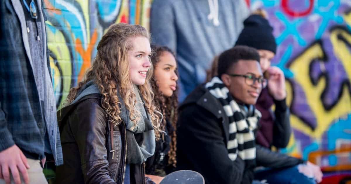 5 diverse teenagers sitting and standing in front of a graffiti wall wearing scarves, flannel shirts, leather jackets, stocking caps