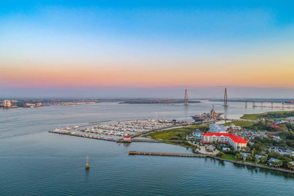 Aerial view of Charleston South Carolina. Harbor and marina are in lower half of the photo on the right side. A single boat is in the water at the bottom of the photo. In the middle, off in the horizon, is a large bridge that spans across the entire photo, joining Charleston to a barrier island. Sky is shades of blue, pink and yellow.