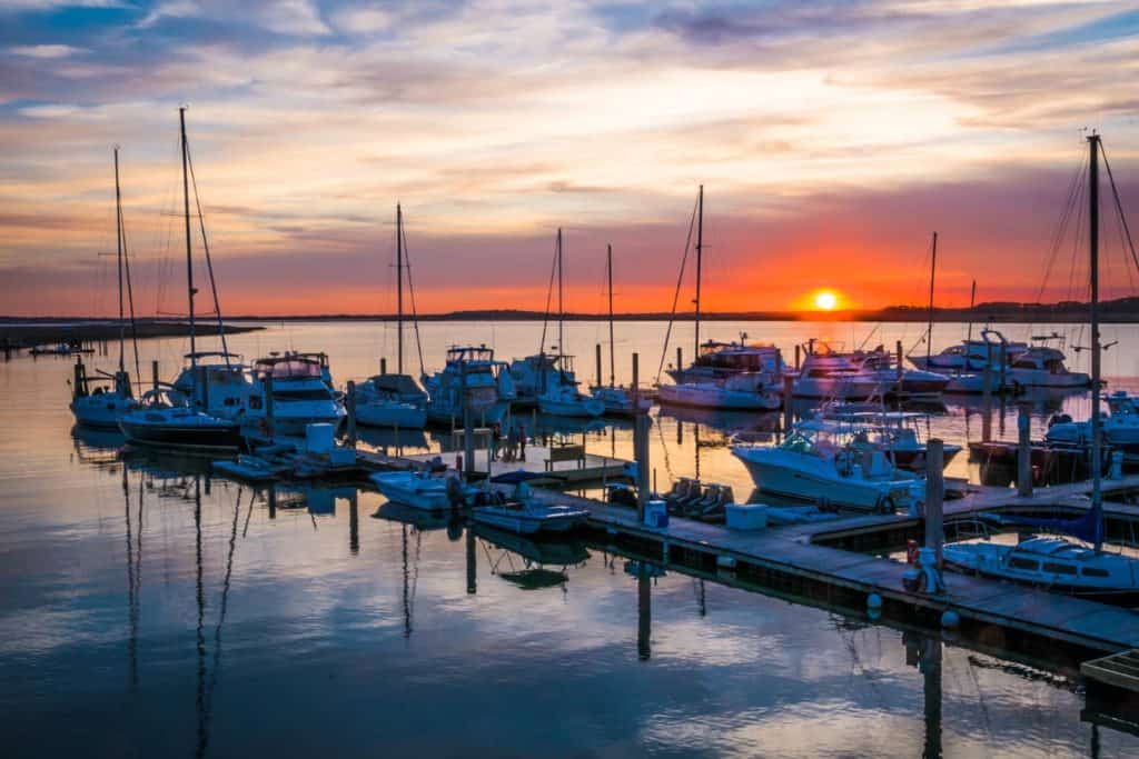 Folly Beach marina. Lots of fishing boats, speed boats and sailboats at Folly Beach. Water is clear and sunset with colors of orange, purple, pink, blue and white in the background.