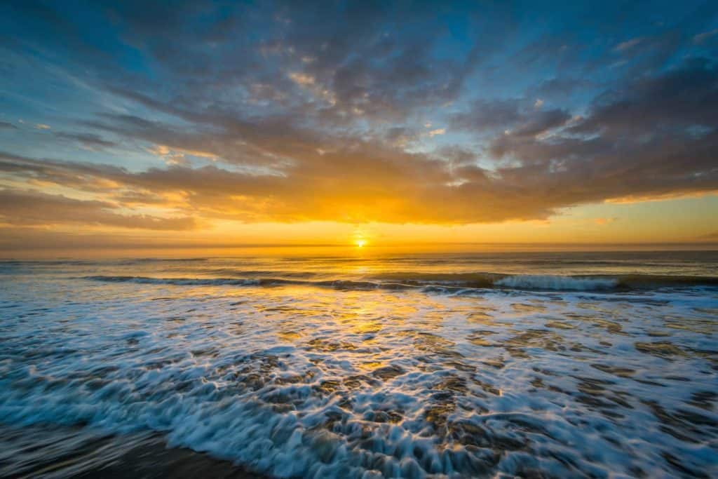 Sunrise at Isle of Palms Beach in South Carolina. Waves crashing on the shore, middle of picture is the horizon with sun just beginning to rise, sky in the center is bright yellow. Top of photo is puffy white clouds and blue skies.