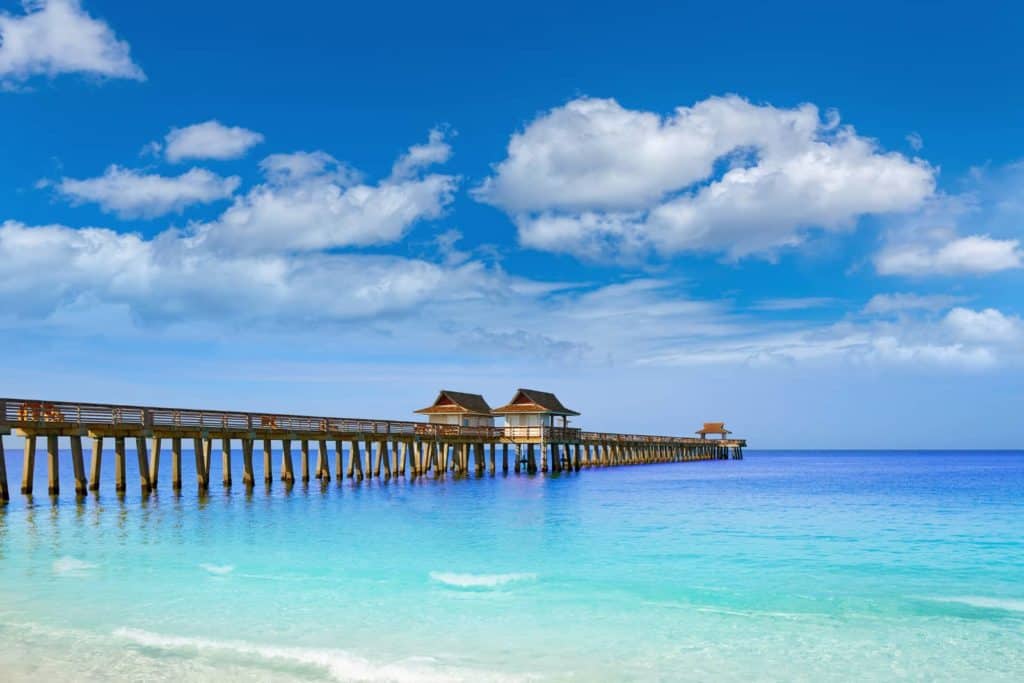 Naples pier jutting into the ocean from the left to the middle of the picture. Bright blue skies and a few puffy white clouds. Clear water that changes from pale green to royal blue.