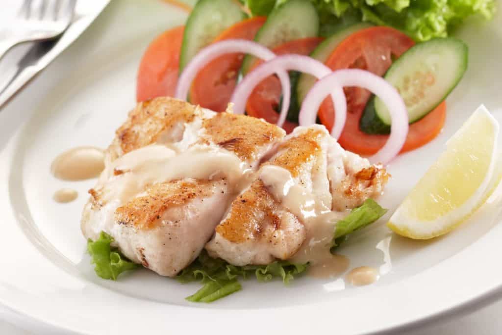 White plate with cooked white fish with a light brown sauce over a bed of lettuce and next to a slice of lemon. In the background is a pretty arrangement of onions, tomatoes and cucumbers.