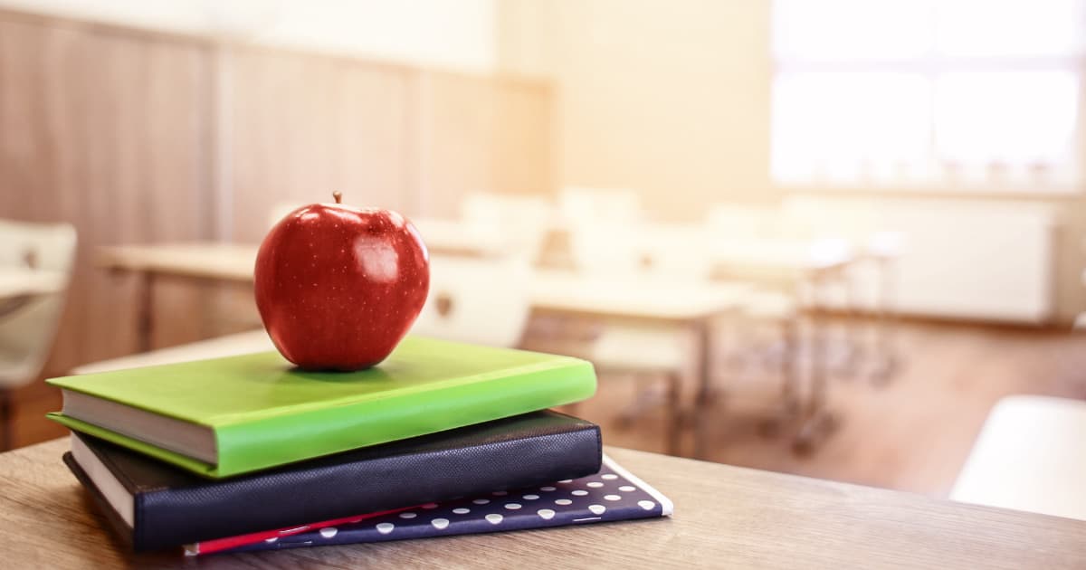 Empty classroom in the background. Stack of blue and green books on a table in the foreground with a bright red apple centered on top of the books. Cover image for retirement gifts for teachers because classroom is empty.