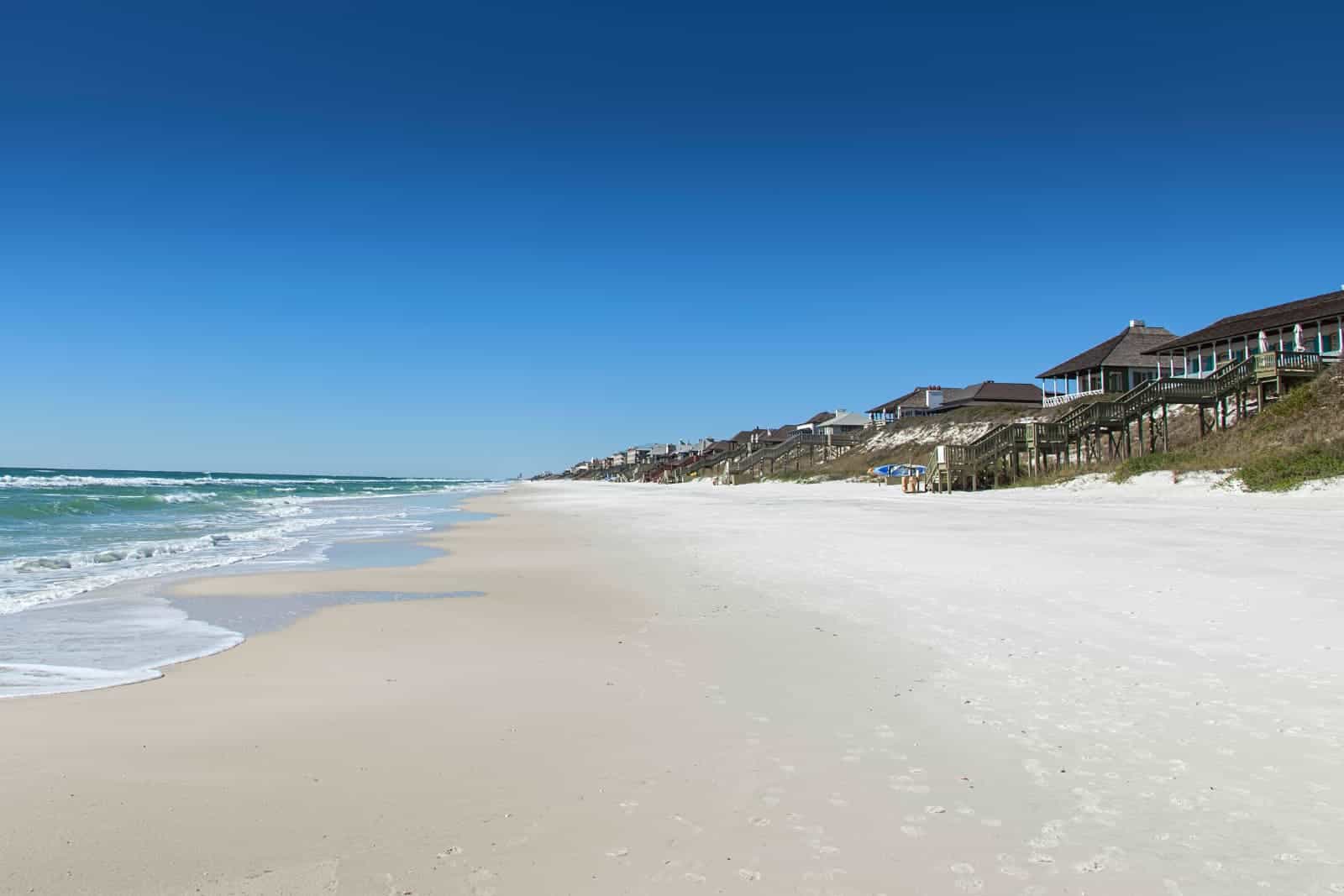 View of Rosemary Beach on 30A Florida with ocean waves on the left, sand in the middle, row of villas on the right. Sky covers top half of the picture and is royal blue.