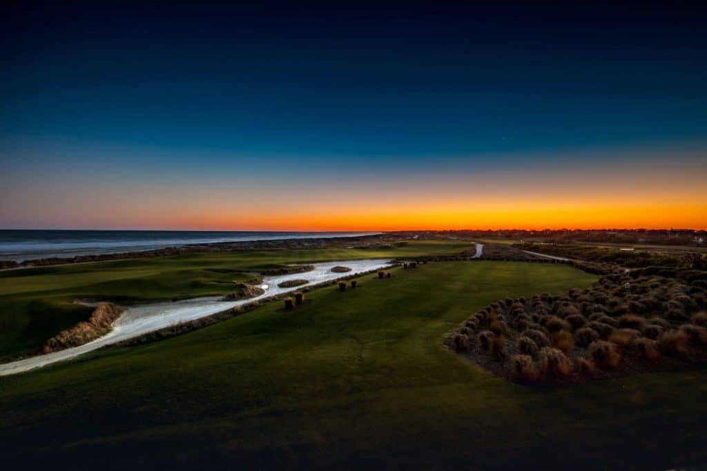 Sunset from the Ryder Cup bar, overlooking Ocean Course on Kiawah Island in South Carolina. Fairway is in the center of the photo, moving diagonally to the right until it fades into the horizon as the sun is setting. On the left are sand traps and sand dunes, then grass until it fades into the ocean. Sky is a deep blue with colors of orange, yellow, red and pink.