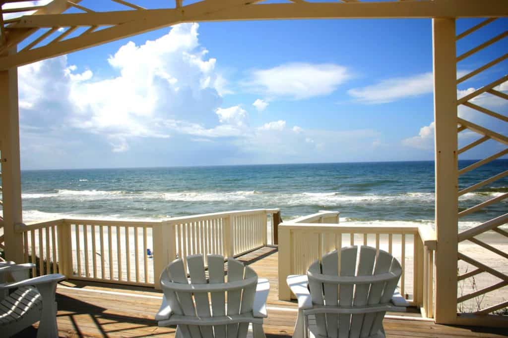 Looking out over the ocean from a large pergola that frames the photo. Two white adirondack chairs are facing a walkway with railings that goes toward the beach. Blue ocean waves, a bright blue sky and puffy white clouds