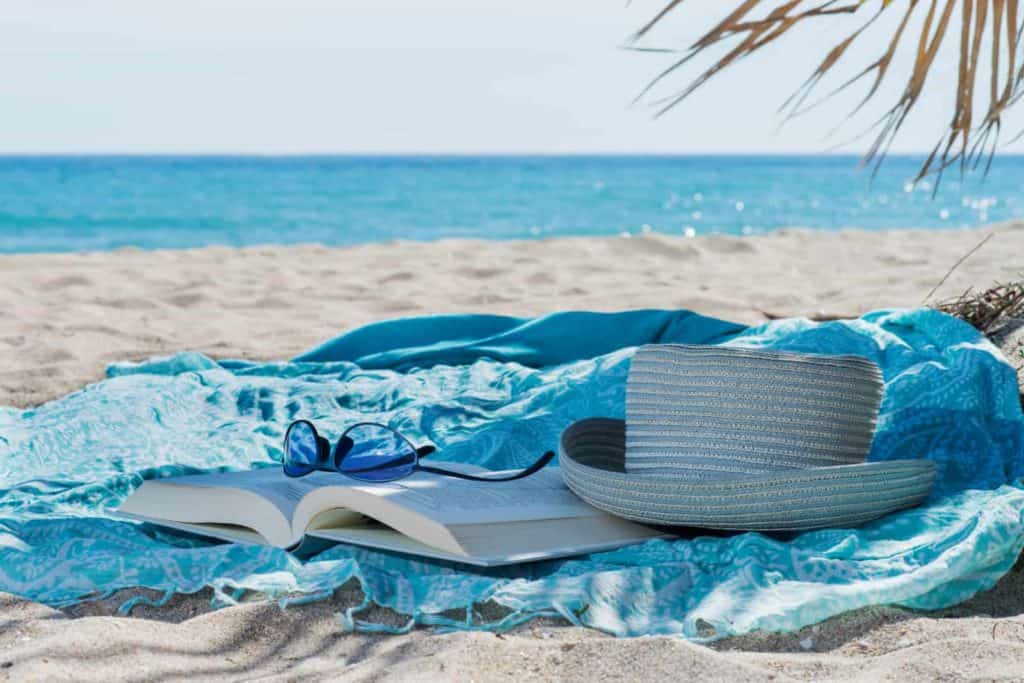 Thin blue beach towel on the sand with bright blue ocean in the background. Palm branch frames right corner of photo. Striped blue straw hat, open hardcover book and blue sunglasses turned upside down on the book. All rest on the blue blanket.