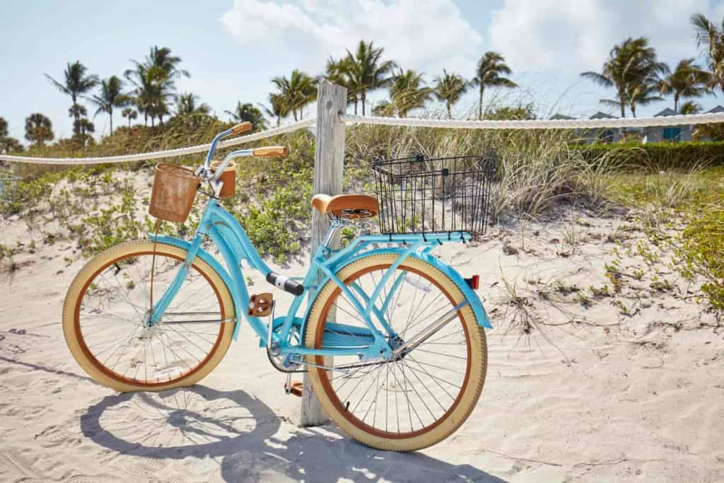 Bright blue bike with tan wheels, seat and handlebars leaning against a wooden post in the sand. Bike has a black basket on the back and a brown basket on the front. Grasses and palm trees are in the background.