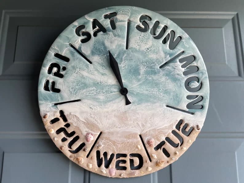 Day of the week clock divided into seven sections with abbreviations of each day around the outer edge of the clock. Painted to look like a beach. Ocean waves at the top, waves crashing to shore in the middle, sandy beach with shells at the bottom