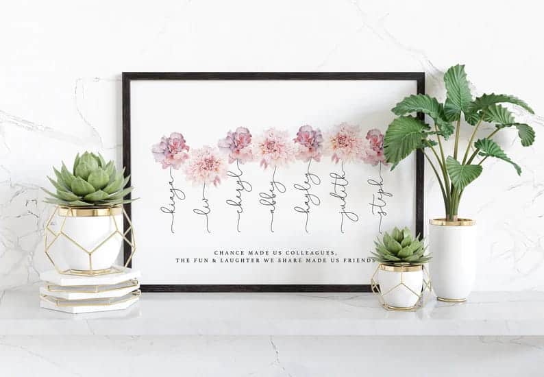 Print with white background in black frame leaning against white marble wall with three plants in foreground. Plants are in white and gold pots. Print has pink and purple flowers with names in cursive as the stems. Words at the bottom in black say "Chance made us colleagues. The fun & laughter we share made us friends."