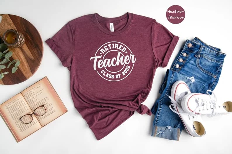Heather maroon tee with white text that says "Retired Teacher class of 2022" on a white background. Surrounded by wood lazy susan with candle, open book with glasses on top, jeans and tennis shoes